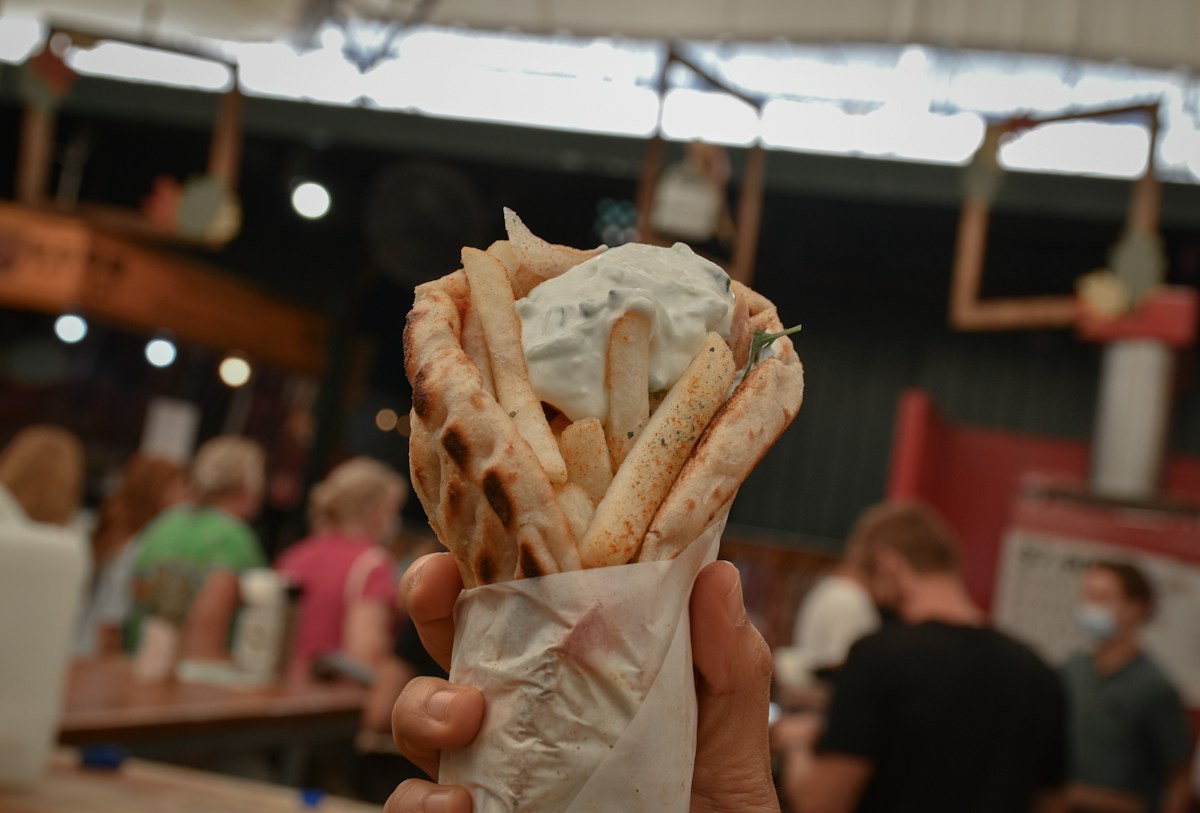 Hand holding a pita gyro filled with fries and topped with tzatziki at a busy food market.
